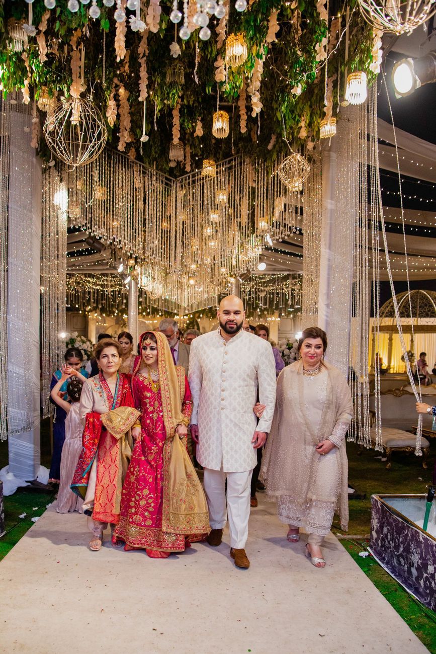 Bride and groom walking together with their mothers beneath a beaded chandelier installation at the reception, Mashal & Danial's wedding