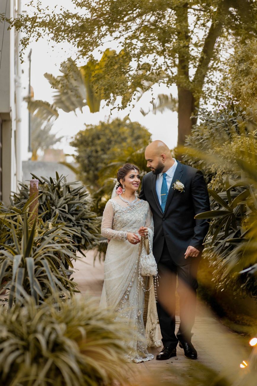 Bride and groom walking together through a garden path, bride in an ivory embellished saree, Mashal & Danial's wedding