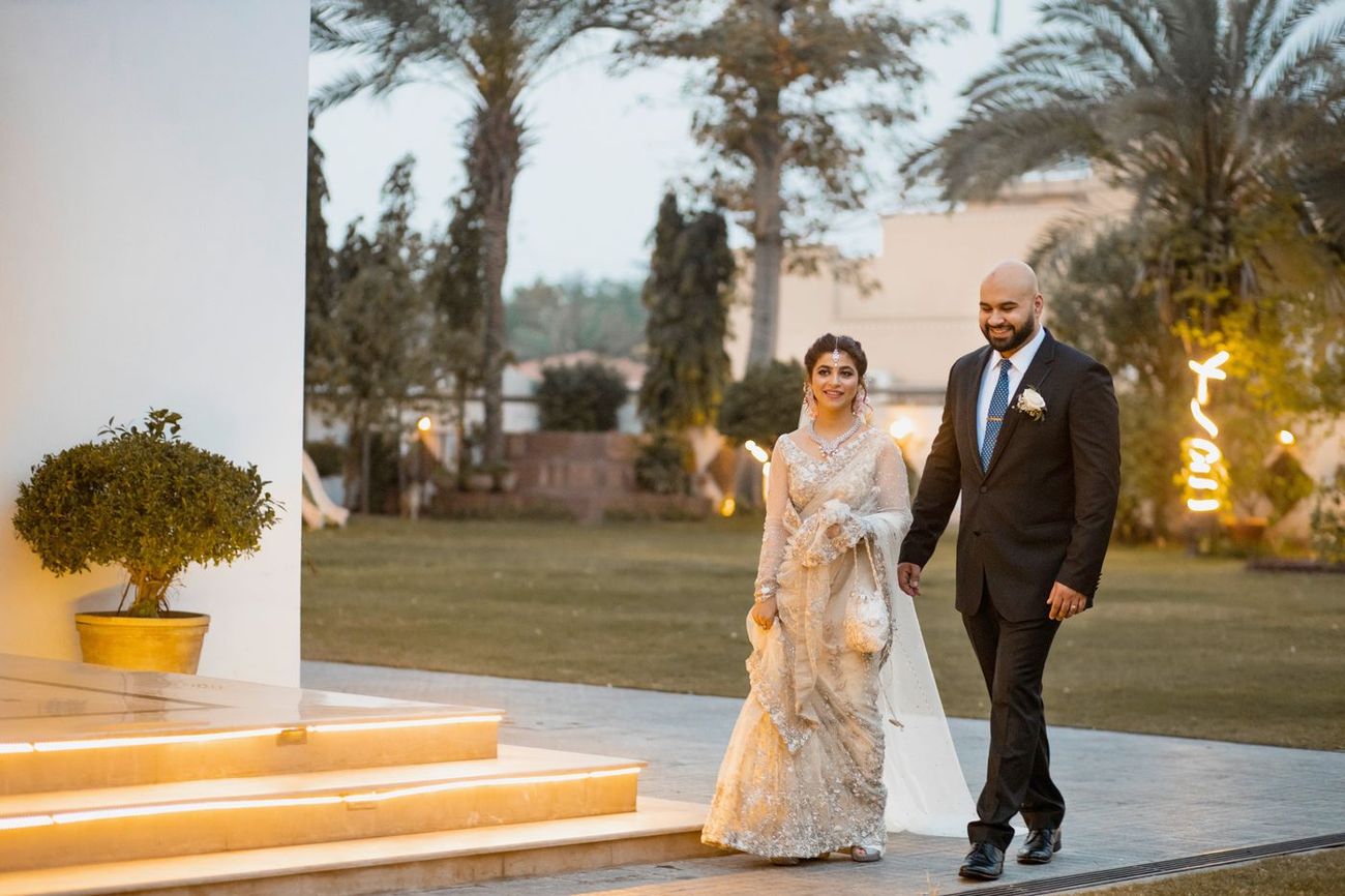 Bride and groom walking together at dusk, a lit-up neon initial sign visible in the garden, Mashal & Danial's wedding