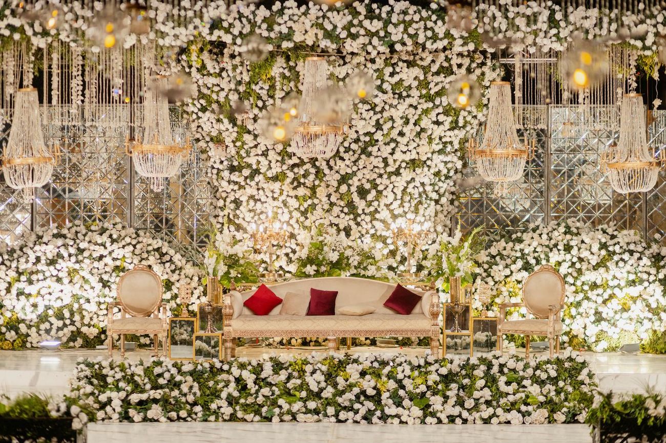 Wide shot of the empty walima stage decorated with a white floral wall and crystal chandeliers, Mashal & Danial's wedding