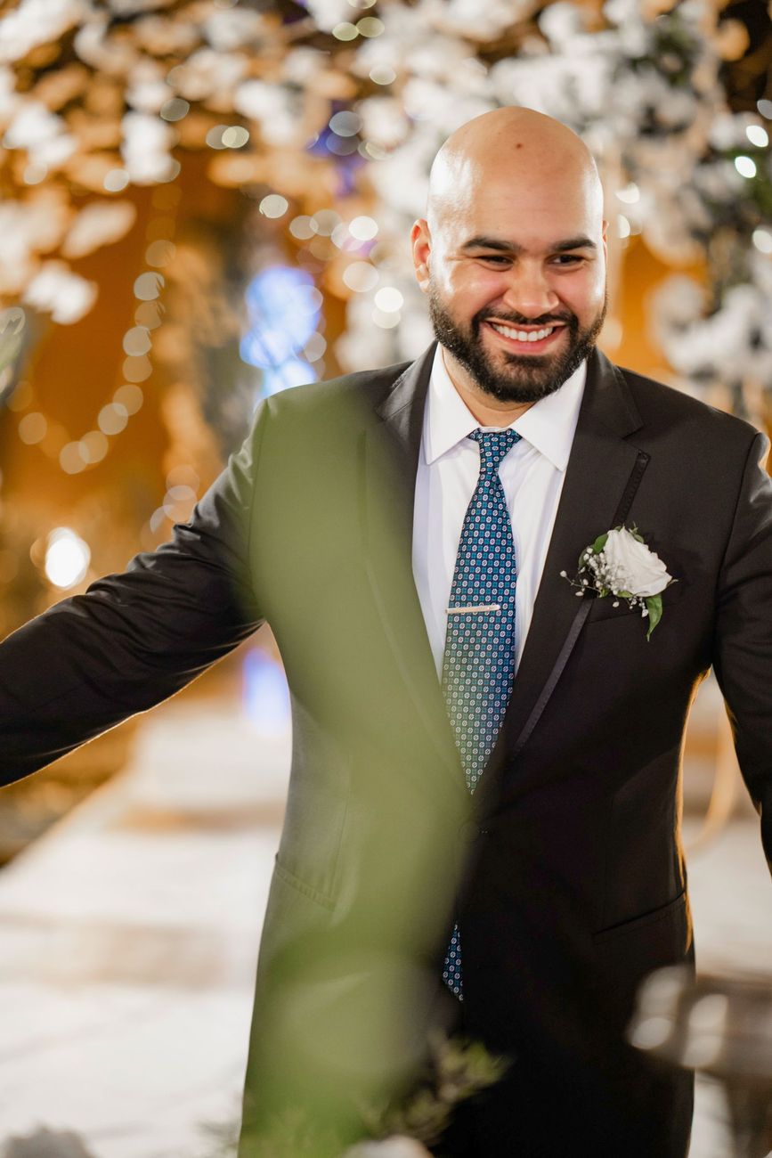 Solo groom portrait smiling at the walima reception, white floral decor blurred behind him, Mashal & Danial's wedding