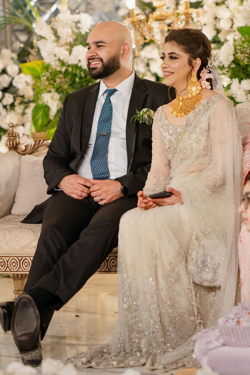 Bride and groom seated together on a sofa at the walima, surrounded by white floral decor, Mashal & Danial's wedding