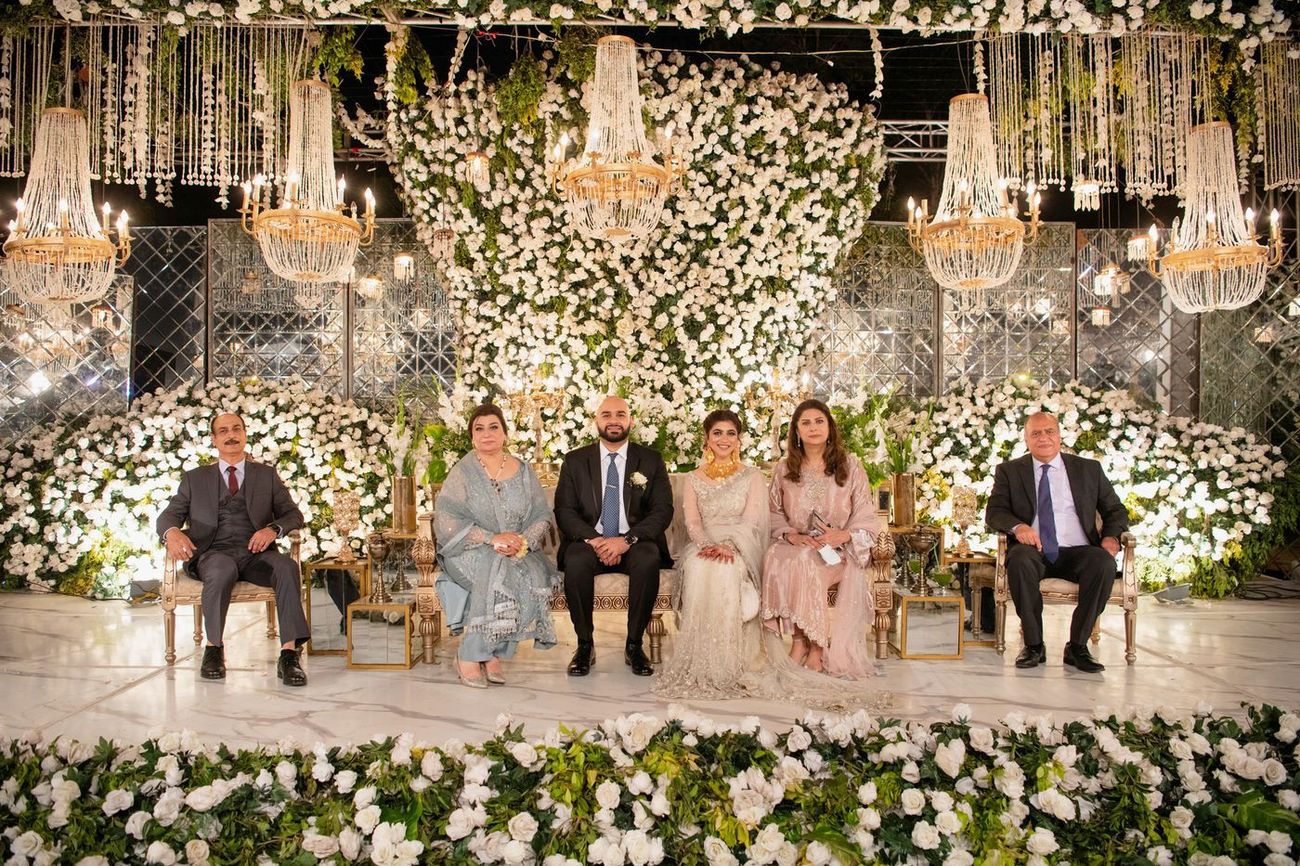 Bride and groom seated with family members at the walima reception, white floral wall behind them, Mashal & Danial's wedding