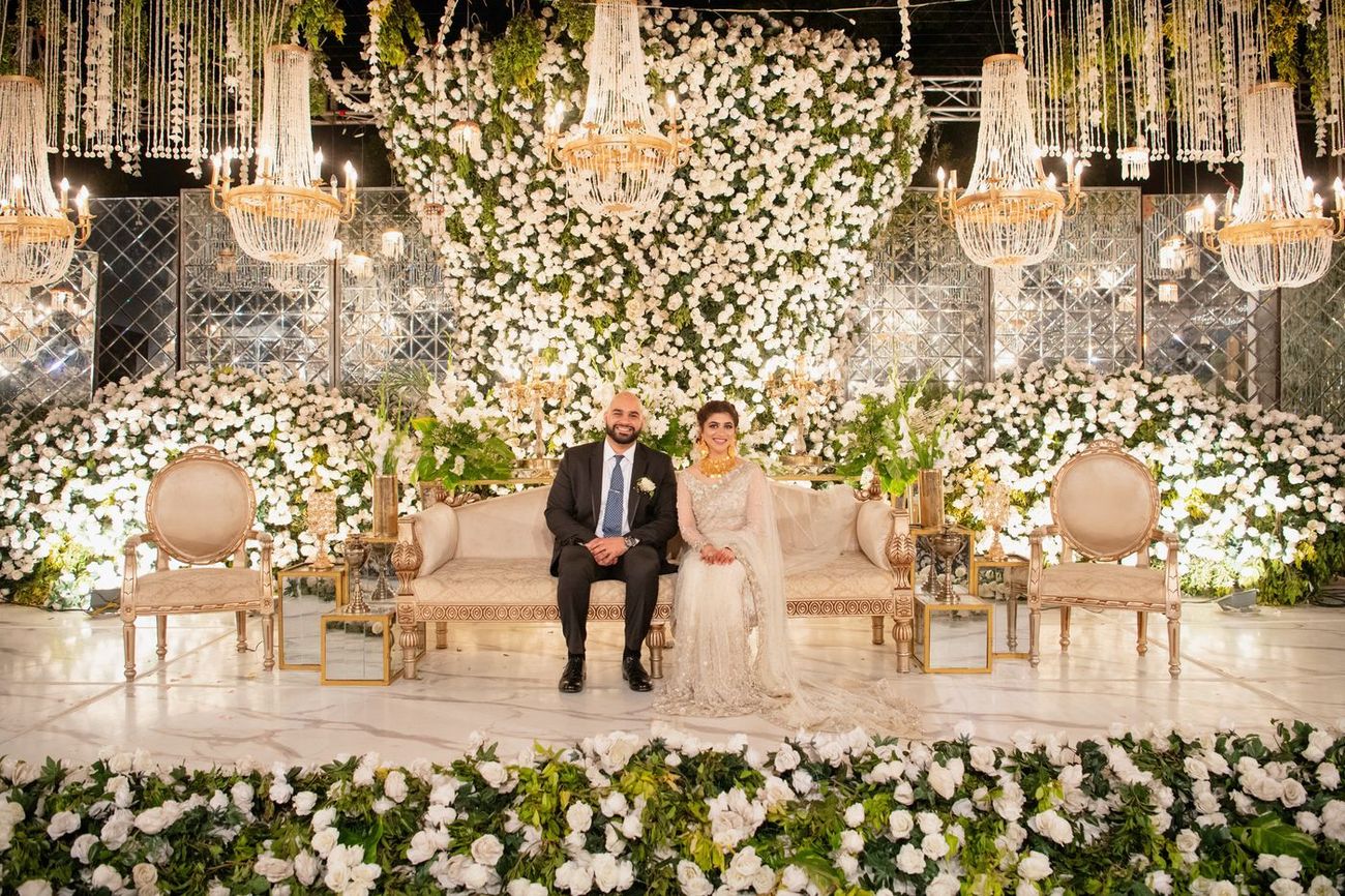 Bride and groom seated together on a sofa at the walima stage, surrounded by white roses, Mashal & Danial's wedding