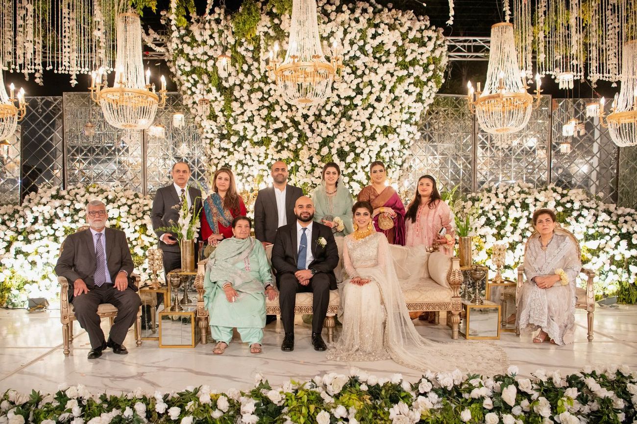 Extended family group photo around the bride and groom at the walima, white floral backdrop behind them, Mashal & Danial's wedding