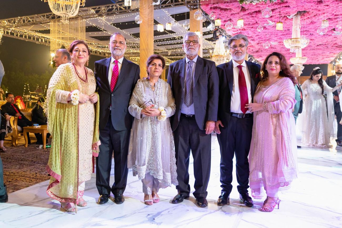 Group of family friends posing together at the walima reception, bride and groom visible in the background, Mashal & Danial's wedding