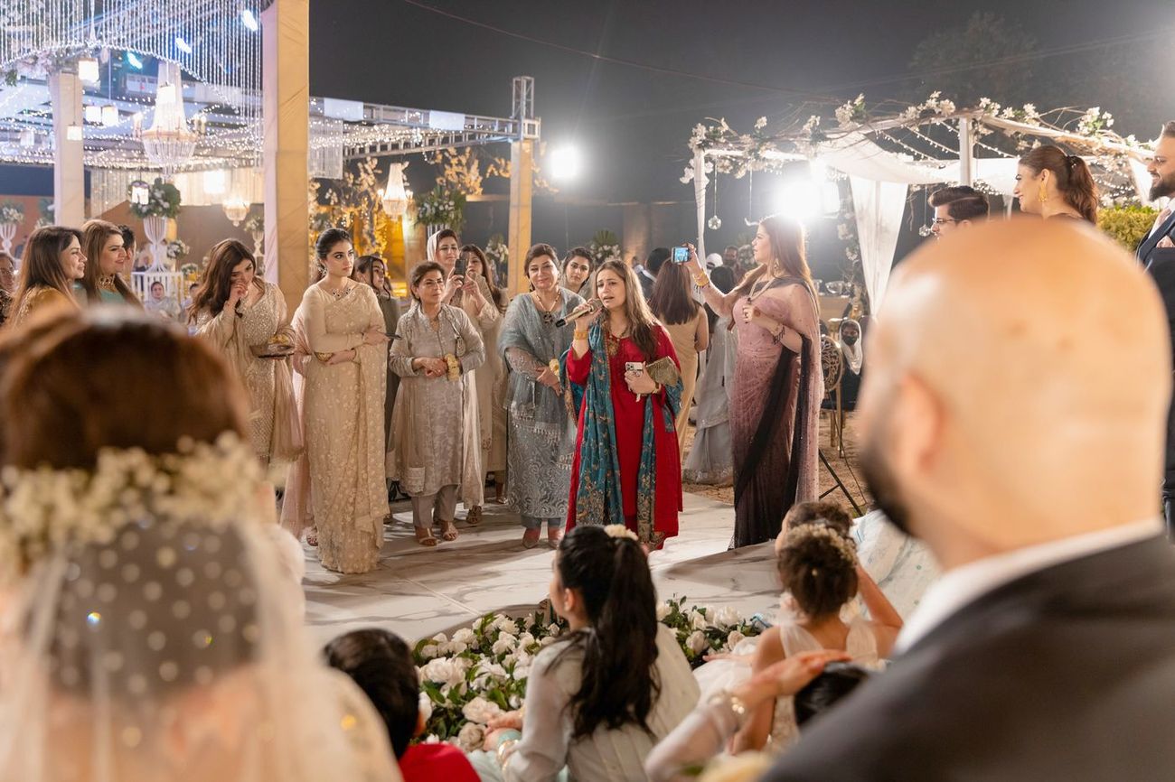 A family member giving a speech with a microphone at the reception as guests gather around, Mashal & Danial's wedding