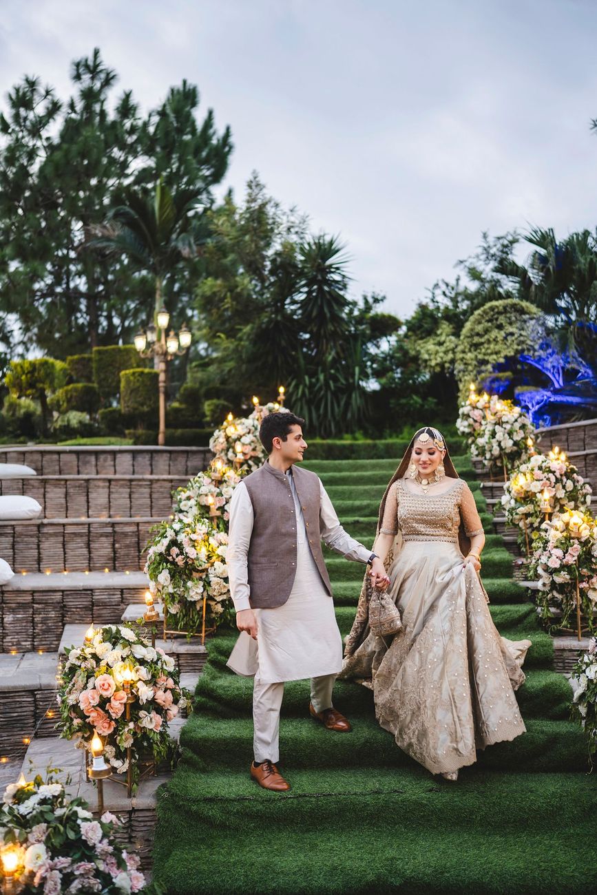 Bride and groom walking down a grass-covered staircase lined with floral arrangements at dusk, wide shot of the full amphitheater venue, Nayha & Rafay's wedding in Islamabad