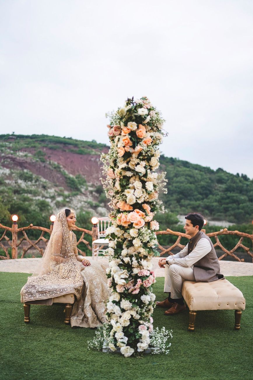 Bride and groom seated facing each other on benches either side of a tall floral installation, hillside backdrop, Nayha & Rafay's wedding in Islamabad