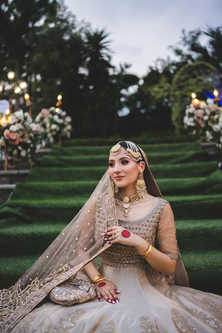 Solo bridal portrait seated on the floral-lined steps at dusk, wearing a gold matha patti and layered necklace, Nayha & Rafay's wedding in Islamabad