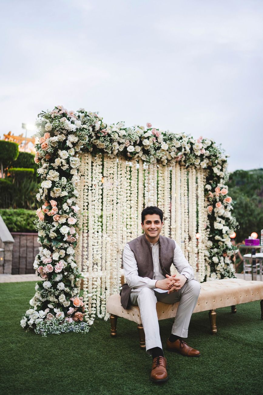 Solo groom portrait seated on a tufted bench in front of a hanging floral and jasmine-strand backdrop, Nayha & Rafay's wedding in Islamabad
