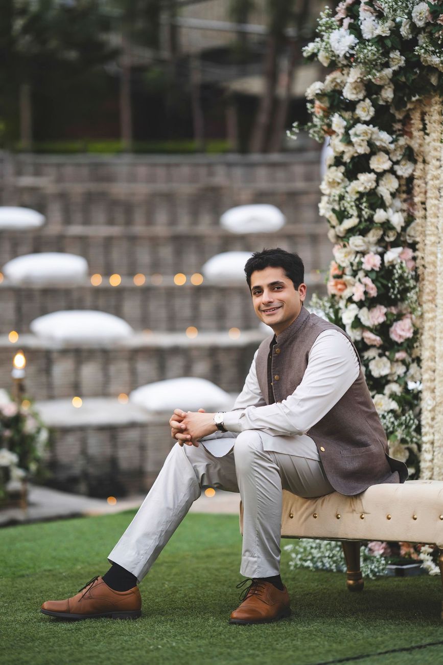 Solo groom portrait seated on a bench in front of floral decor, smiling at the camera, Nayha & Rafay's wedding in Islamabad