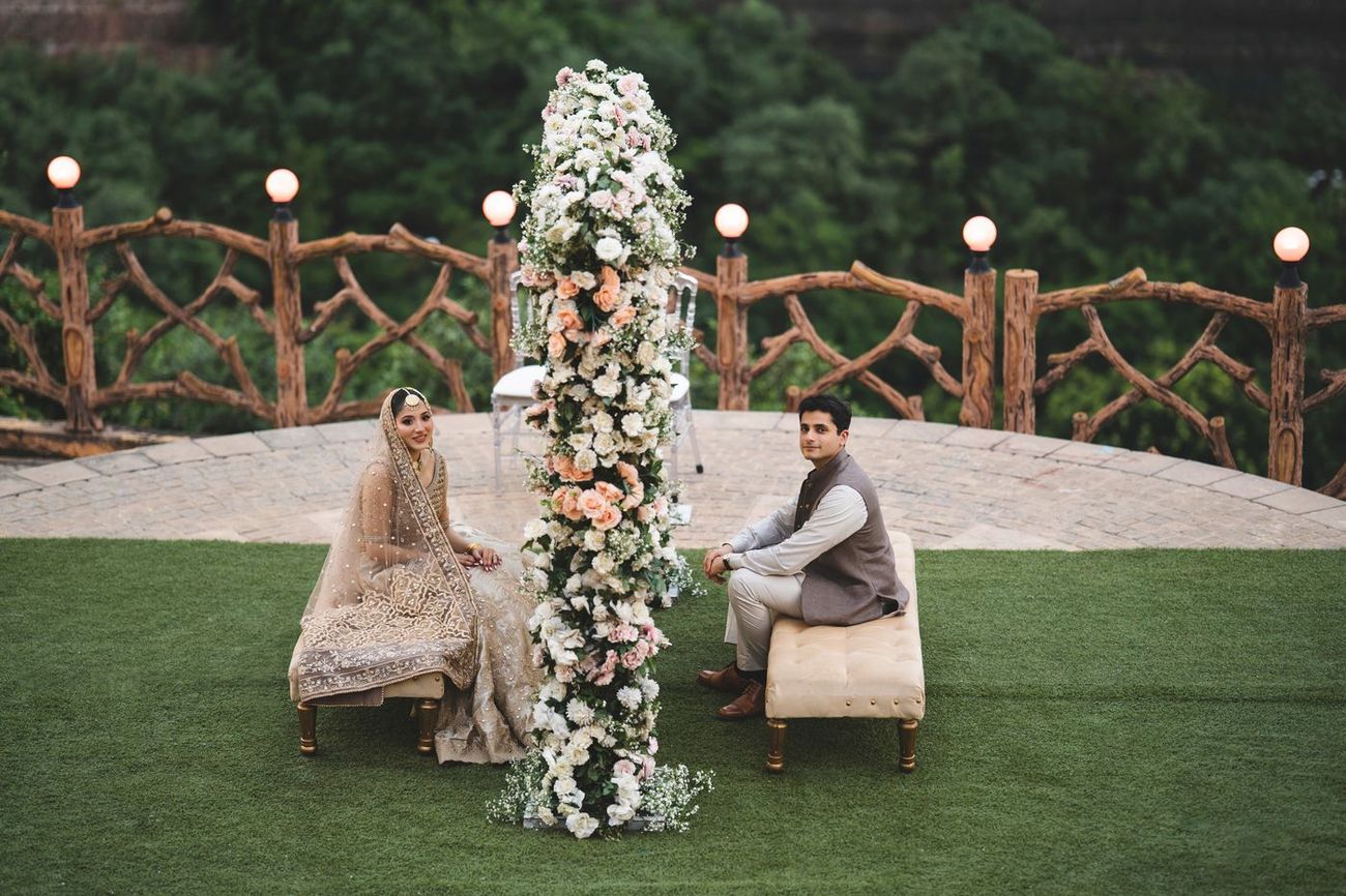 Bride and groom seated on facing benches beside the tall floral installation, both looking toward the camera, Nayha & Rafay's wedding in Islamabad