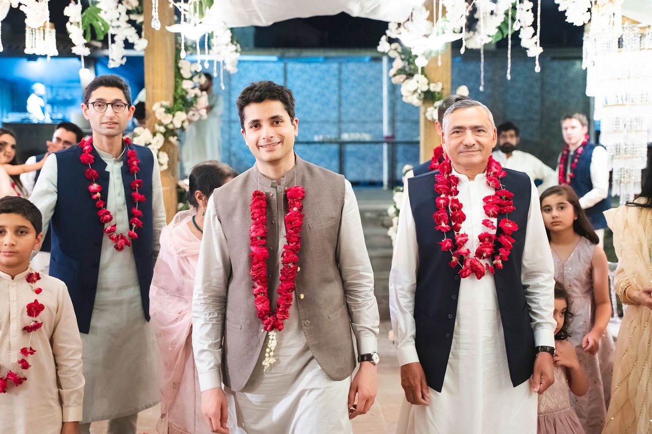 Groom arriving with family members, all wearing red rose garlands, under a floral entrance decor, Nayha & Rafay's wedding in Islamabad