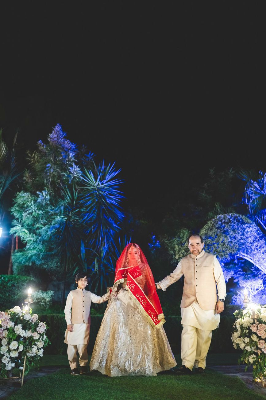 Bride's grand entrance under a red veil, escorted by family through a blue-lit garden at night, Nayha & Rafay's wedding in Islamabad