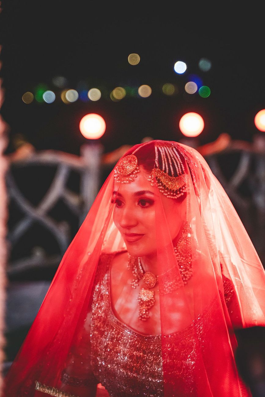 Close-up portrait of the bride under her red veil during her entrance, string lights glowing behind her, Nayha & Rafay's wedding in Islamabad