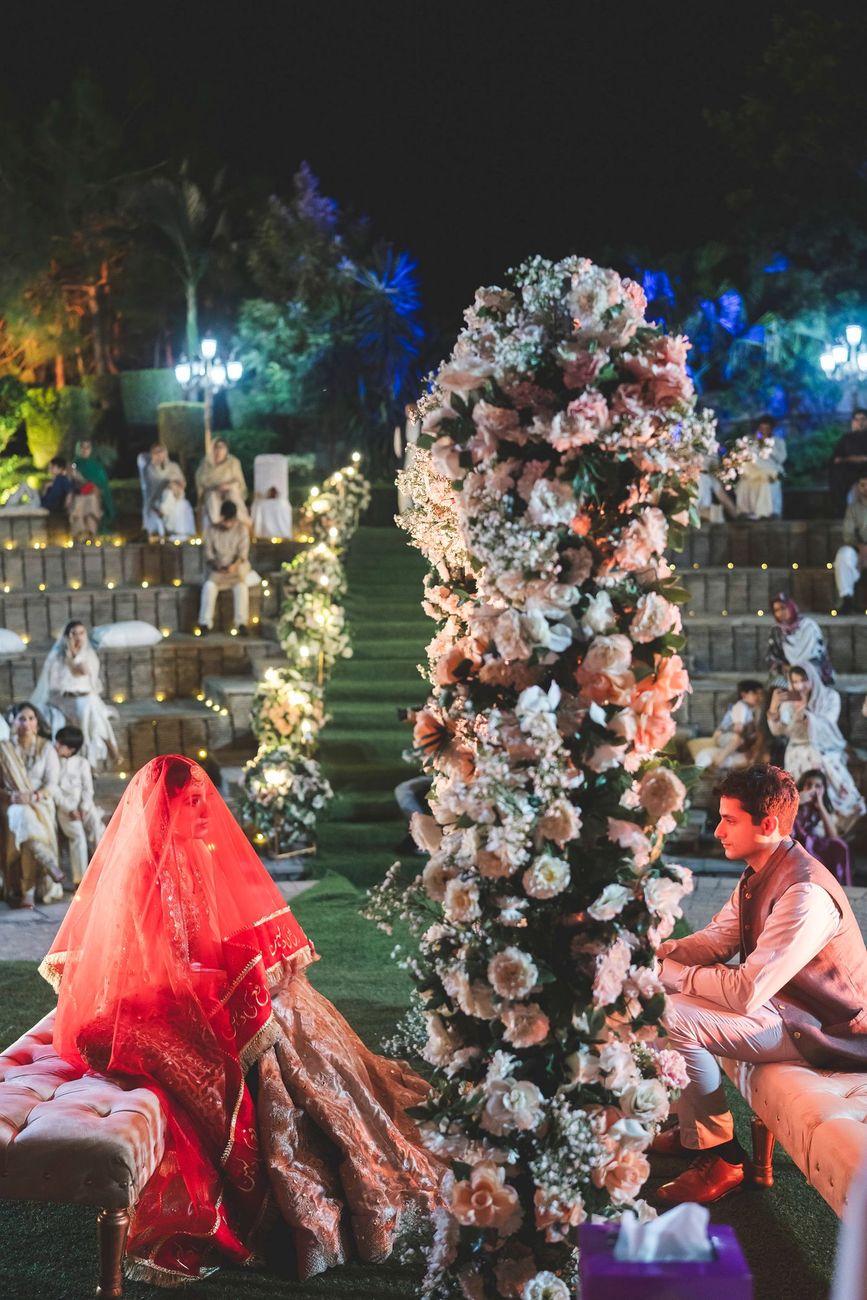 Wide shot of the nikah ceremony, bride seated under her red veil facing the groom across a floral pillar, guests seated in the amphitheater, Nayha & Rafay's wedding in Islamabad