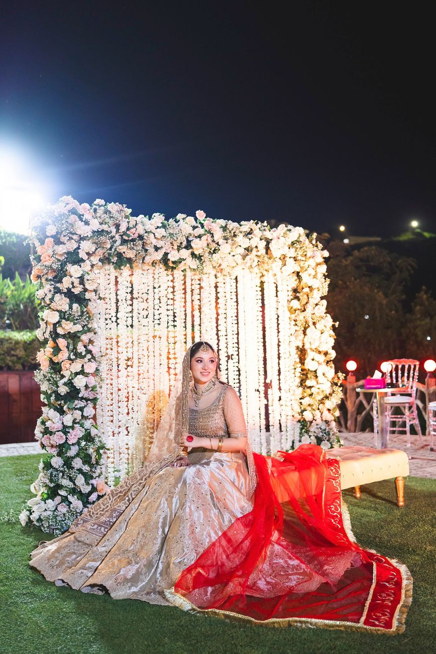 Solo bridal portrait seated in front of the floral curtain backdrop, her red dupatta with Urdu calligraphy spread across the bench, Nayha & Rafay's wedding in Islamabad