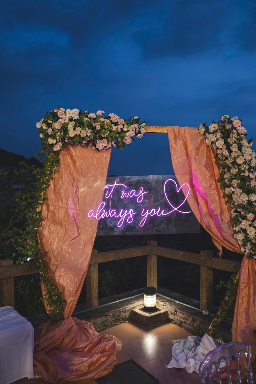 Detail shot of a neon sign reading "It was always you" framed by a floral archway with peach drapes, Nayha & Rafay's wedding decor in Islamabad