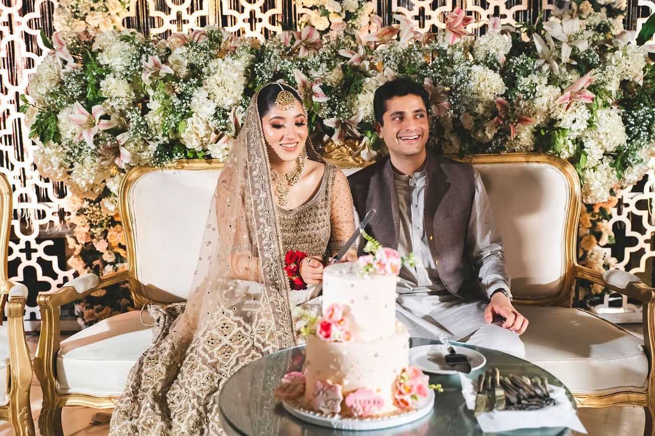 Bride and groom cutting their wedding cake together, seated on a gold-framed loveseat in front of a floral wall, Nayha & Rafay's wedding in Islamabad