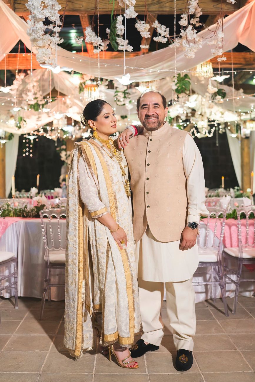Bride's parents posing together at the reception beneath a draped floral ceiling, dining tables visible behind them, Nayha & Rafay's wedding in Islamabad