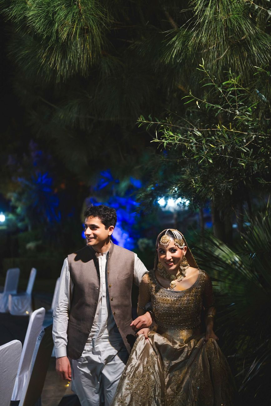 Bride and groom walking together at night beneath pine branches, blue garden lighting in the background, Nayha & Rafay's wedding in Islamabad