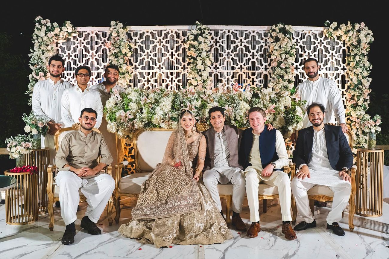 Group portrait of bride, groom, and groomsmen seated together on gold-framed chairs in front of a floral wall, Nayha & Rafay's wedding in Islamabad