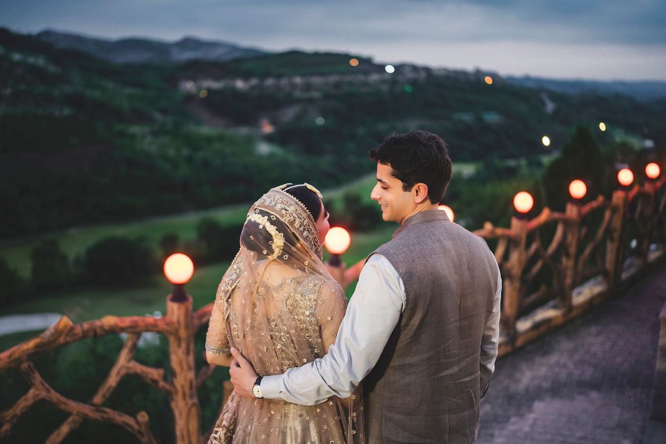 Bride and groom viewed from behind, looking out over the hillside view from a wooden-fenced terrace at dusk, Nayha & Rafay's wedding in Islamabad