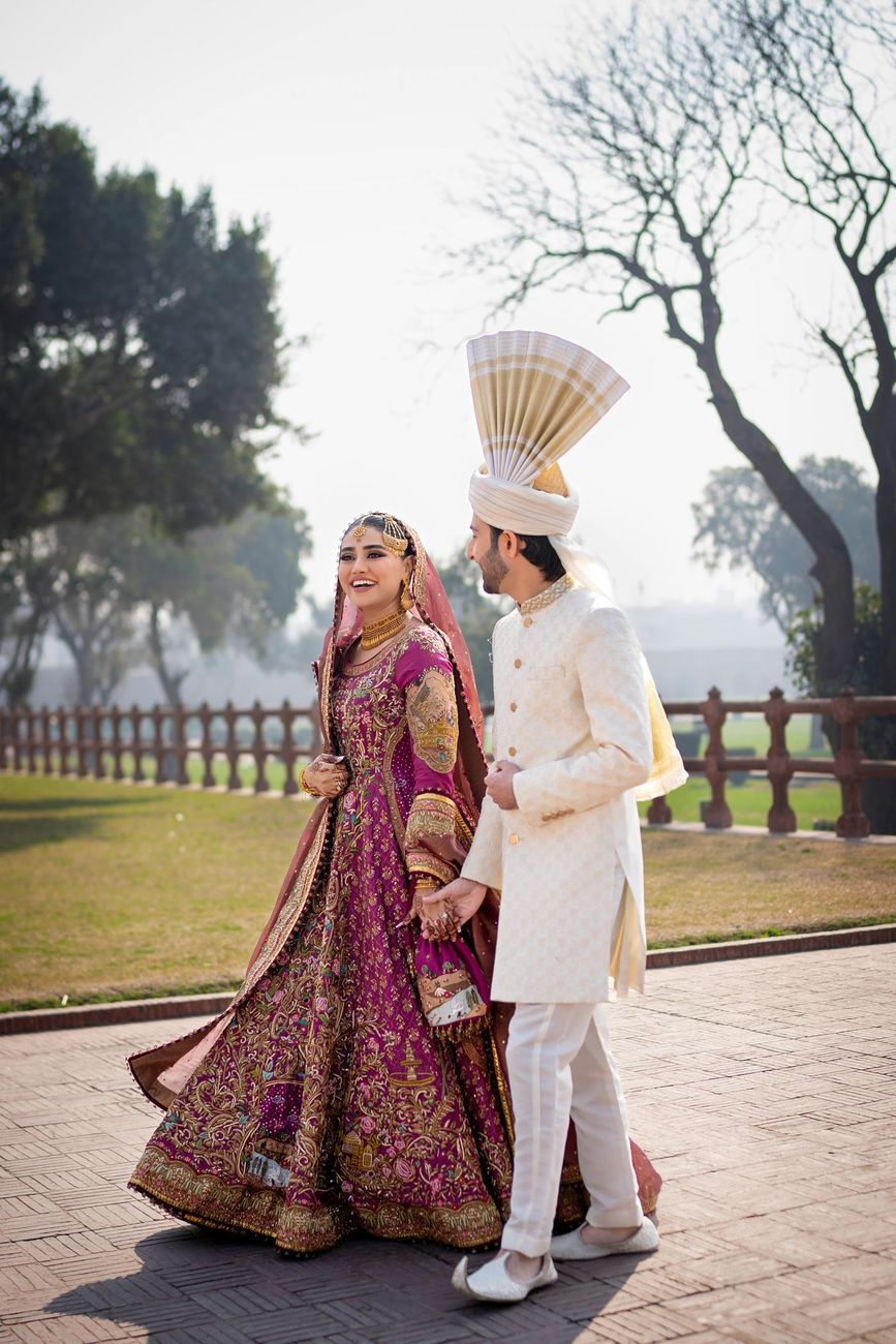 Bride and groom walking hand in hand along a brick garden path, her in a magenta and gold embroidered lehenga, him in an ivory sherwani with a tall pankha turban fan, Samar & Anoosh's wedding at Lahore Fort
