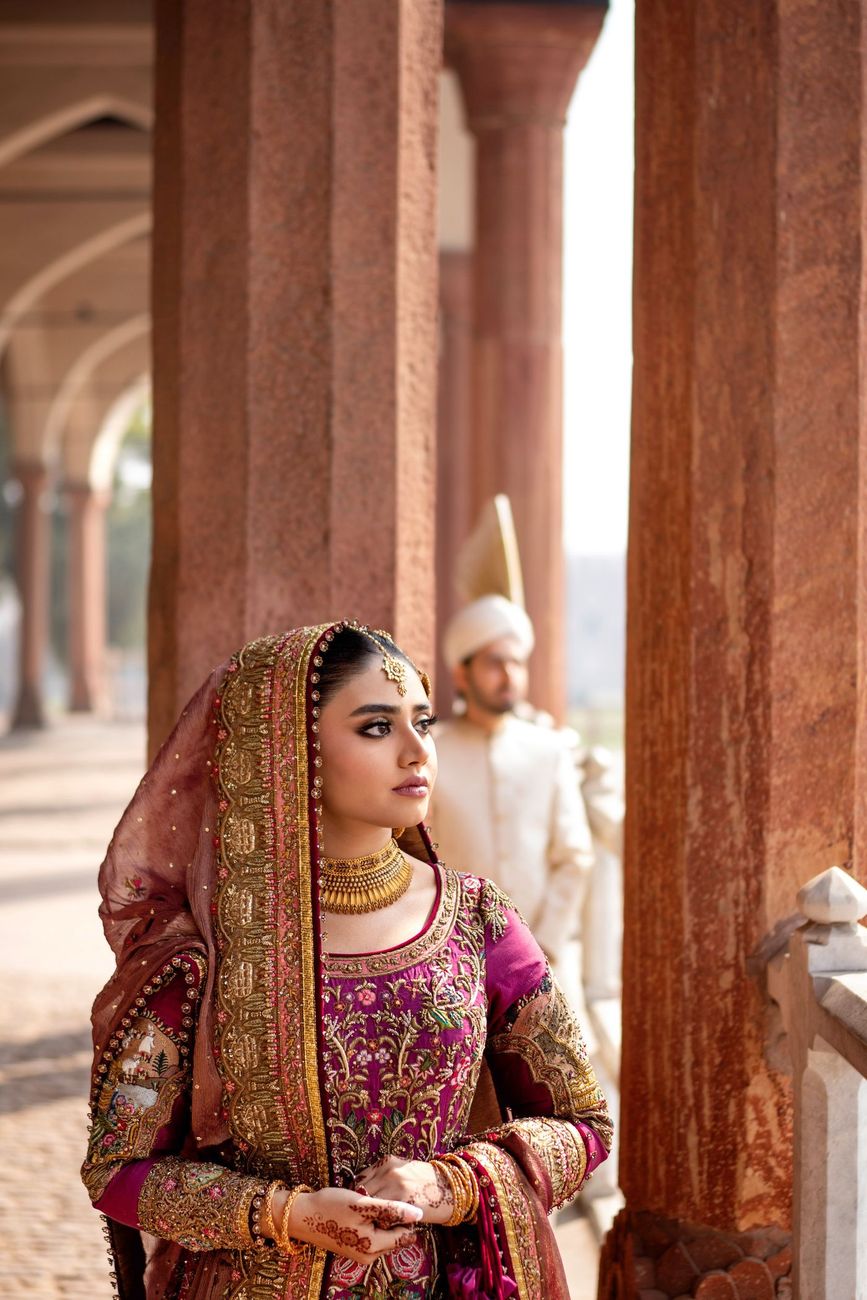 Close-up bridal portrait beneath the fort's red sandstone colonnade, looking off to the side, groom blurred in the background, Samar & Anoosh's wedding at Lahore Fort