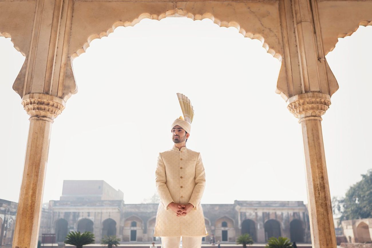 Solo groom portrait standing beneath a scalloped sandstone archway, hands clasped, the fort's courtyard visible behind him, Samar & Anoosh's wedding at Lahore Fort