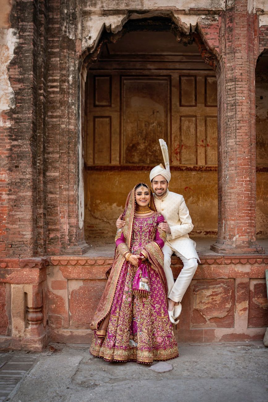 Bride and groom posed together in a restored brick archway, groom seated on the ledge with his arm around her, Samar & Anoosh's wedding at Lahore Fort