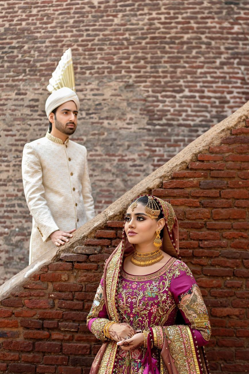 Bride in the foreground wearing a matha patti and gold jewelry, groom standing on the steps behind her against a brick wall, Samar & Anoosh's wedding at Lahore Fort