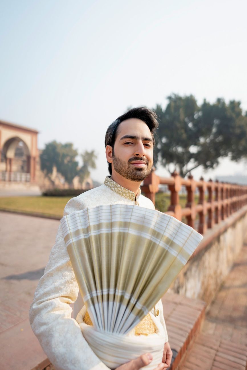 Close-up groom portrait holding his pankha turban fan, a garden walkway and red sandstone railing blurred behind him, Samar & Anoosh's wedding at Lahore Fort