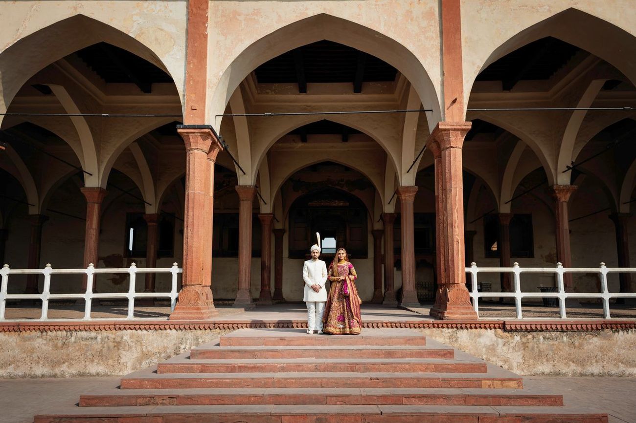 Wide shot of bride and groom standing together beneath the fort's arched colonnade at the top of a stone staircase, Samar & Anoosh's wedding at Lahore Fort