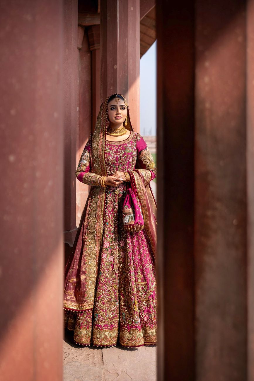 Solo bridal portrait framed between weathered sandstone pillars, holding her potli bag, Samar & Anoosh's wedding at Lahore Fort