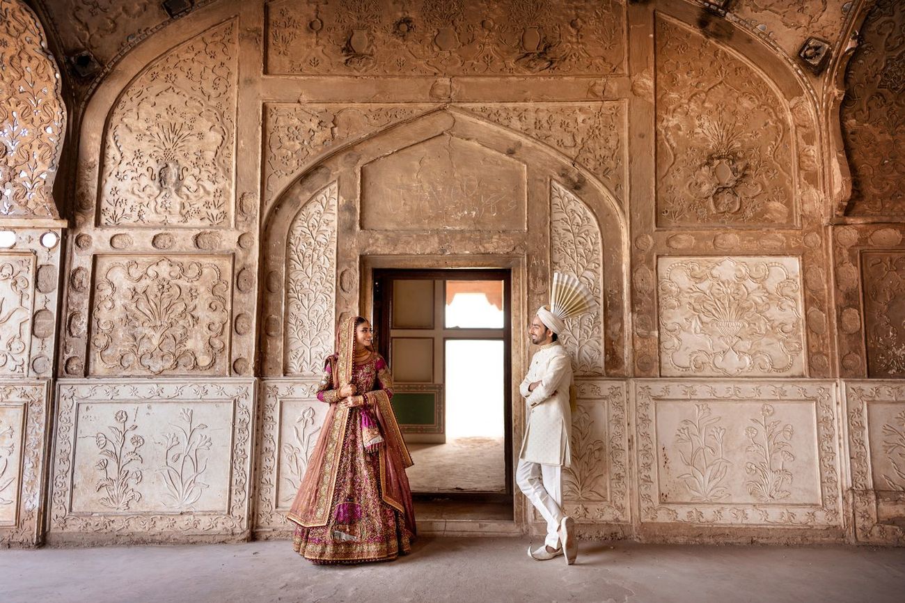 Bride and groom standing near a carved doorway beneath ornately decorated Mughal-era stone archways, exchanging a glance, Samar & Anoosh's wedding at Lahore Fort