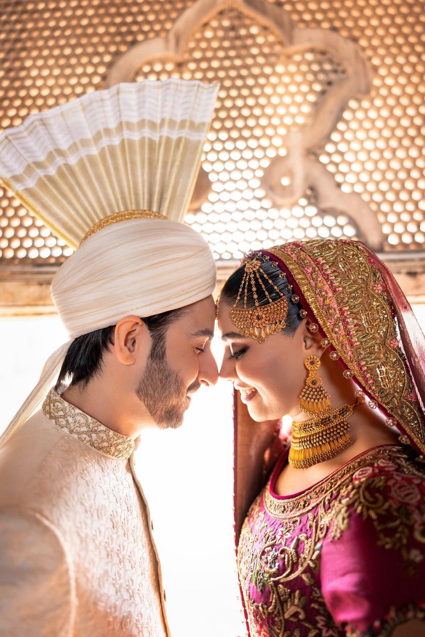 Close-up of bride and groom touching foreheads in front of a sunlit carved jali screen, her gold jhoomar and earrings catching the light, Samar & Anoosh's wedding at Lahore Fort