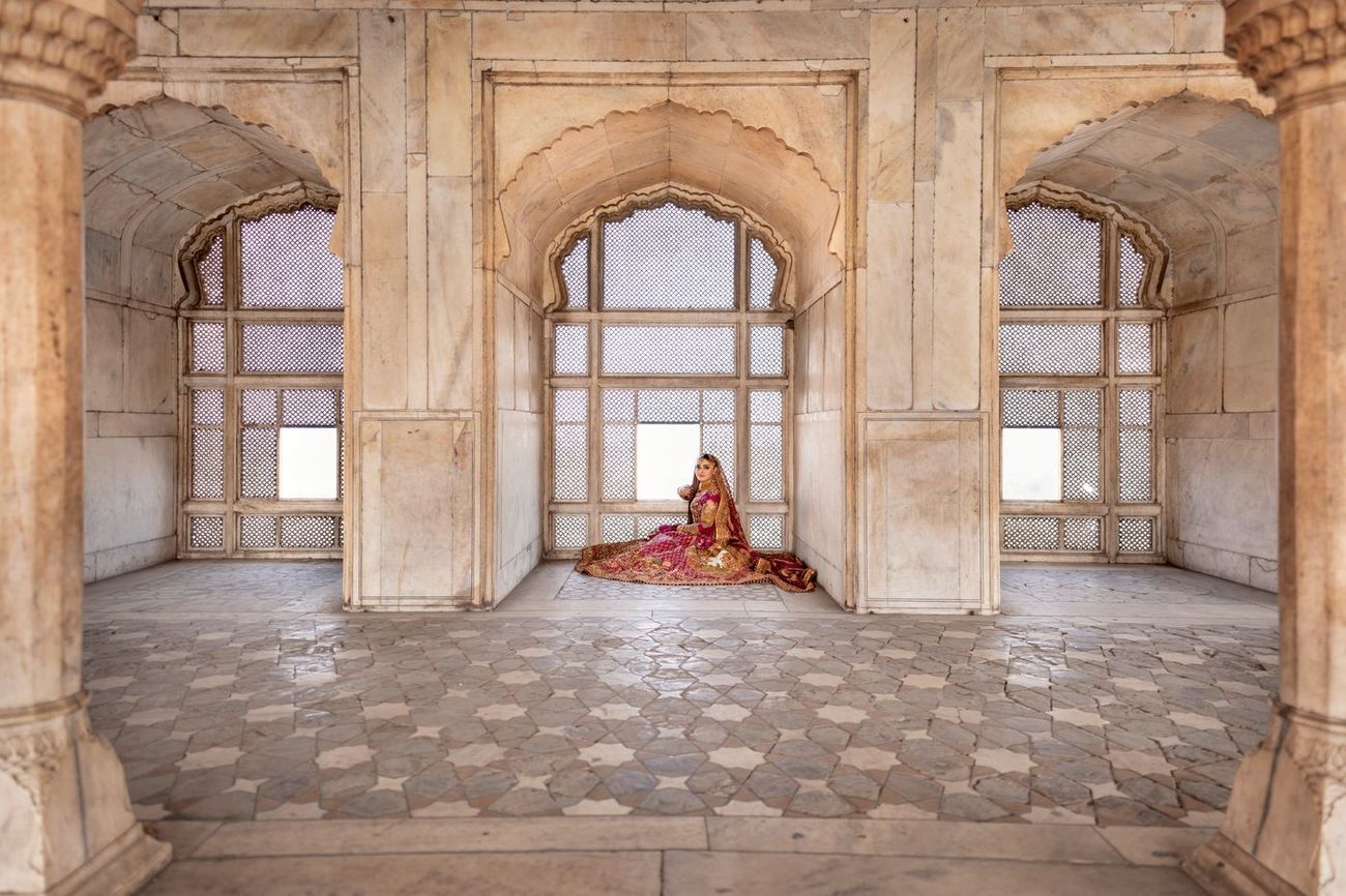 Wide architectural shot of the bride seated alone in a carved marble window alcove, her lehenga pooled across the tiled floor, Samar & Anoosh's wedding at Lahore Fort