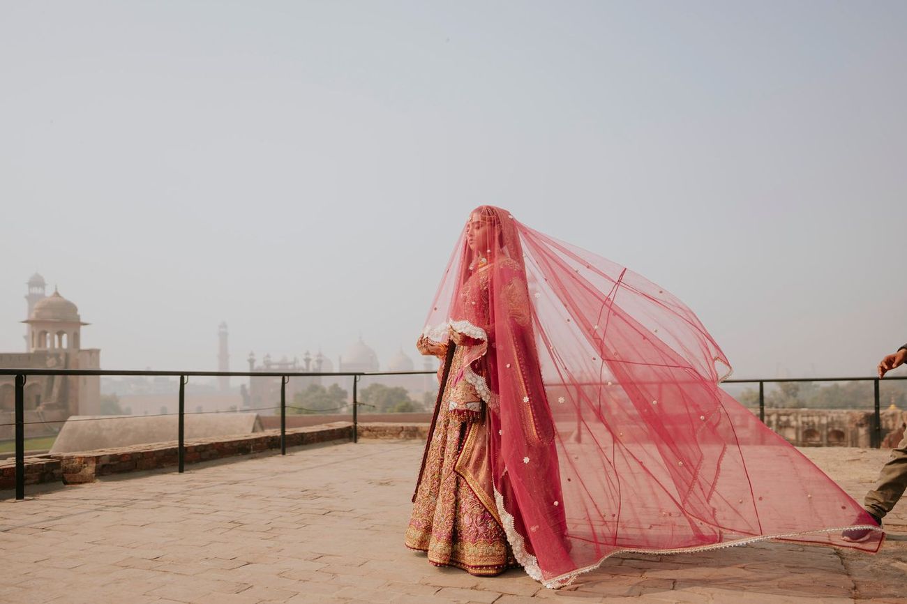 Bride standing on the fort's rooftop terrace, her maroon dupatta billowing dramatically in the wind, Badshahi Mosque's domes visible on the skyline behind her, Samar & Anoosh's wedding at Lahore Fort