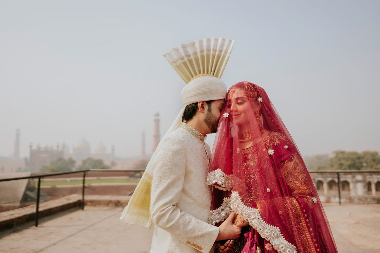 Groom kissing the bride's forehead on the fort's rooftop terrace, Badshahi Mosque visible in the hazy skyline behind them, Samar & Anoosh's wedding at Lahore Fort