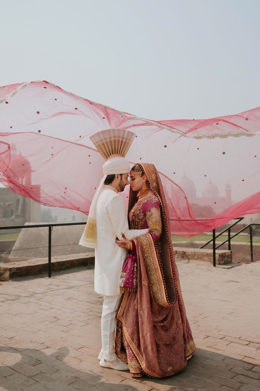 Bride and groom embracing forehead to forehead on the rooftop terrace, her maroon dupatta billowing like a wave behind them with the mosque skyline in the distance, Samar & Anoosh's wedding at Lahore Fort