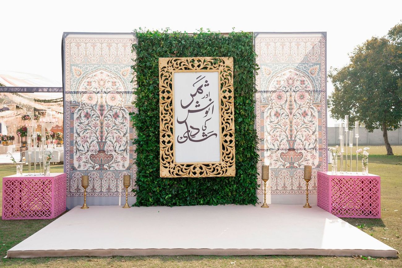 Wedding stage backdrop with a greenery wall, framed Urdu calligraphy signage, and pink Moroccan-style side tables with gold candle stands, Samar & Anoosh's wedding reception
