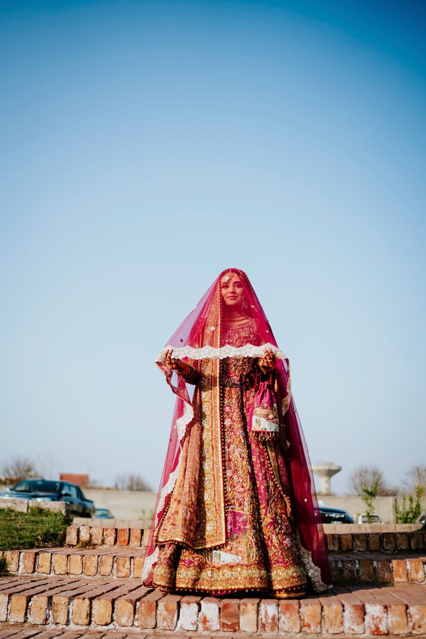 Solo bridal portrait standing on a brick platform against a clear blue sky, her maroon dupatta held open, Samar & Anoosh's wedding