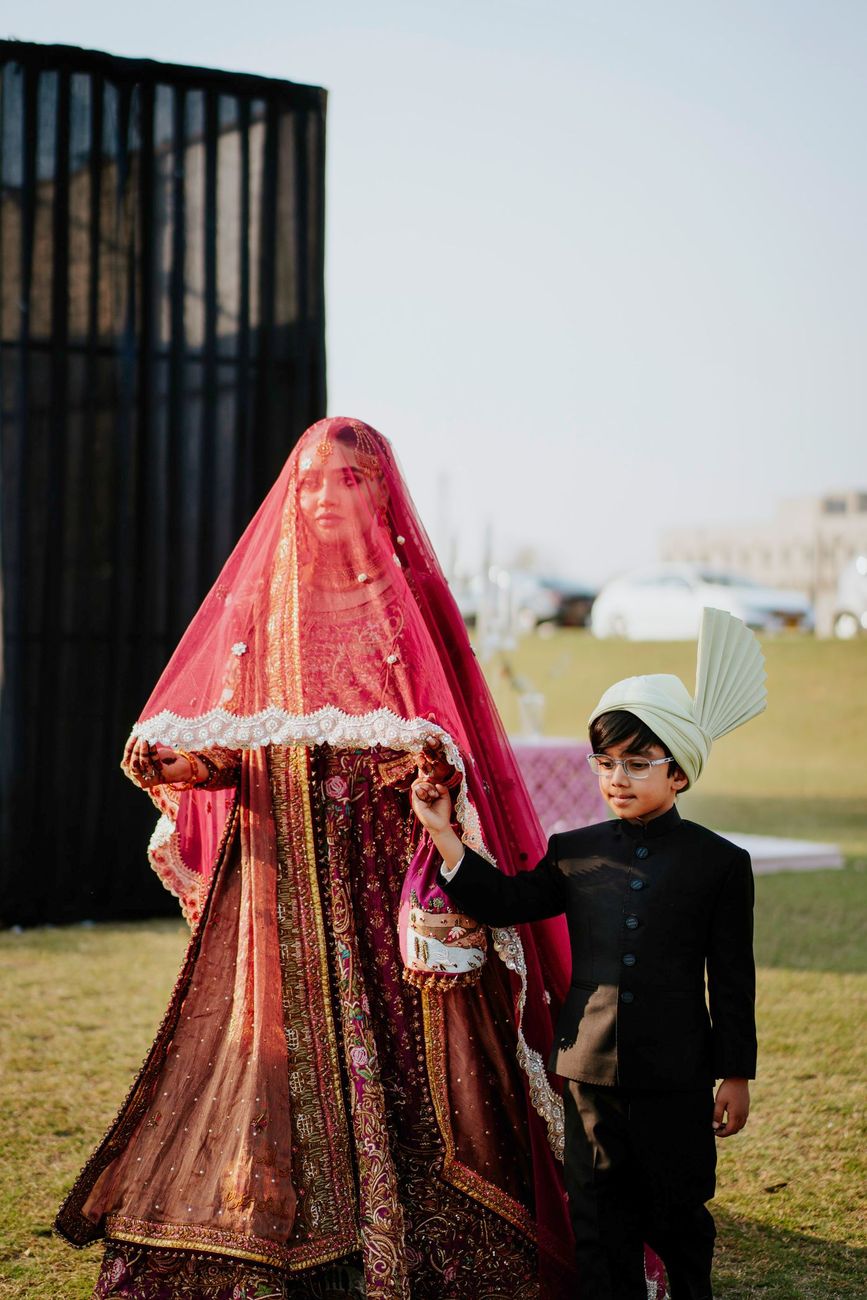Bride veiled in a red dupatta being led by hand by a young boy in a black sherwani and turban, walking across the lawn at the wedding venue