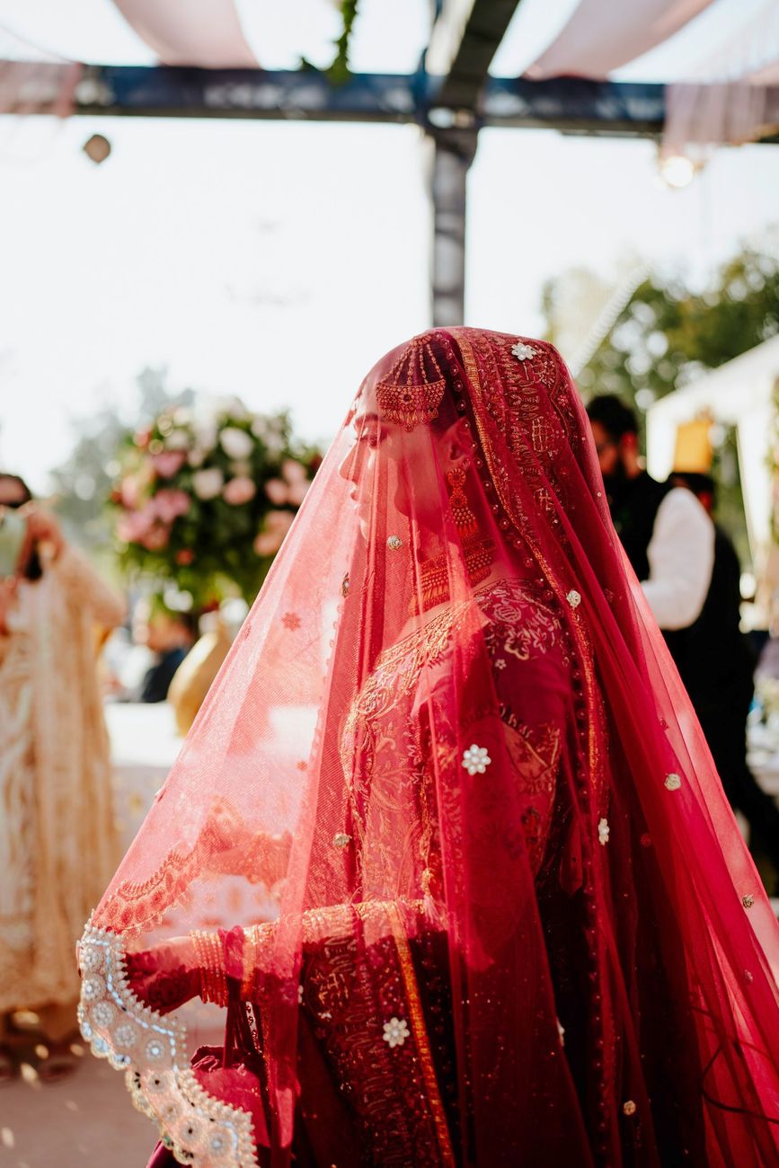 Bride's face veiled beneath a sheer red dupatta, walking past floral arrangements at the wedding venue