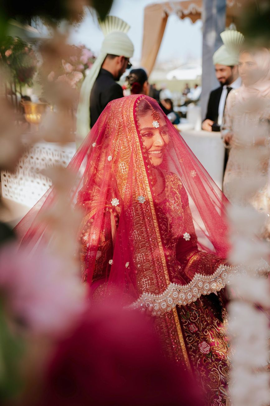 Bride smiling beneath her red veil, framed through pink floral decor at the wedding venue