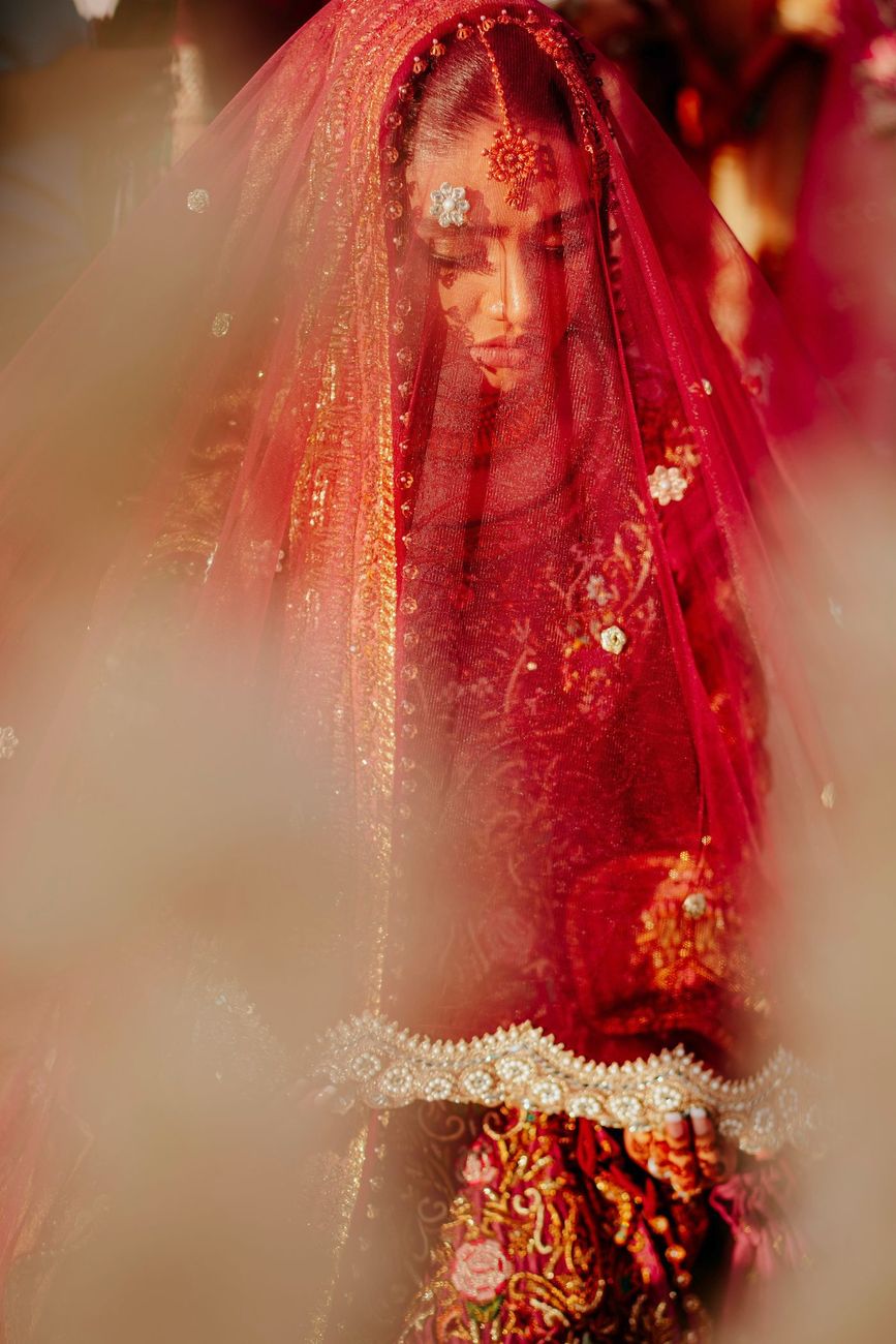 Close-up of the veiled bride framed through dried floral arrangements, her face softly visible beneath the sheer red dupatta