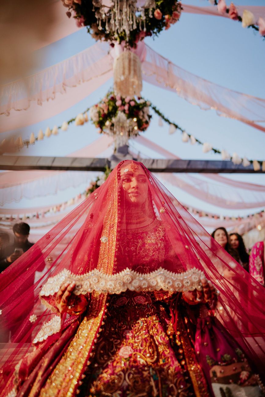 Bride walking beneath the draped pink tent canopy and hanging floral chandeliers, holding the edges of her veil and looking directly at the camera