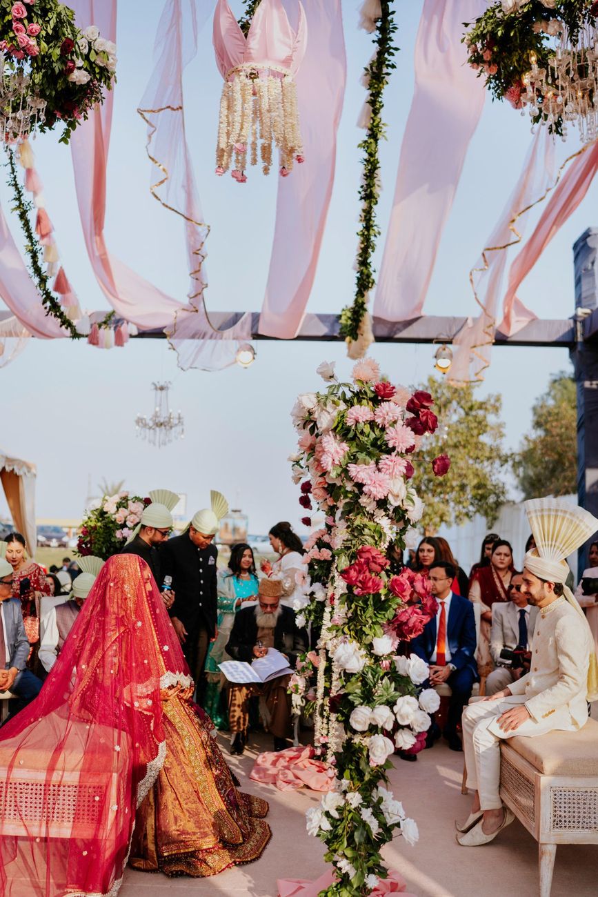 Wide shot of the nikah ceremony underway, groom seated on a bench beside the officiant as the veiled bride kneels facing him, guests seated under the floral-and-drape canopy